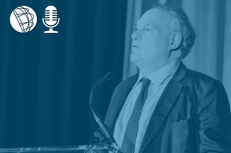 [ai] A man speaking at a podium during an event, with a microphone in front of him. The image is edited in a blue monochrome style. Icons of a globe and a microphone are visible in the top left corner.